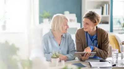 Eine ältere Dame sitzt neben einer jüngeren Frau in Arztkleidung und bekommt auf einem Tablet etwas erklärt