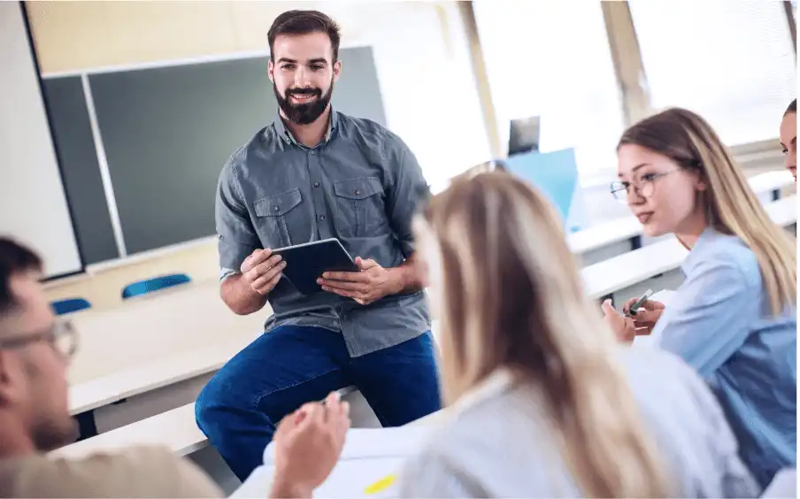 A man sits on a table in a classroom with a chalkboard on the back wall. He is holding an iPad, while four younger people are discussing in the foreground