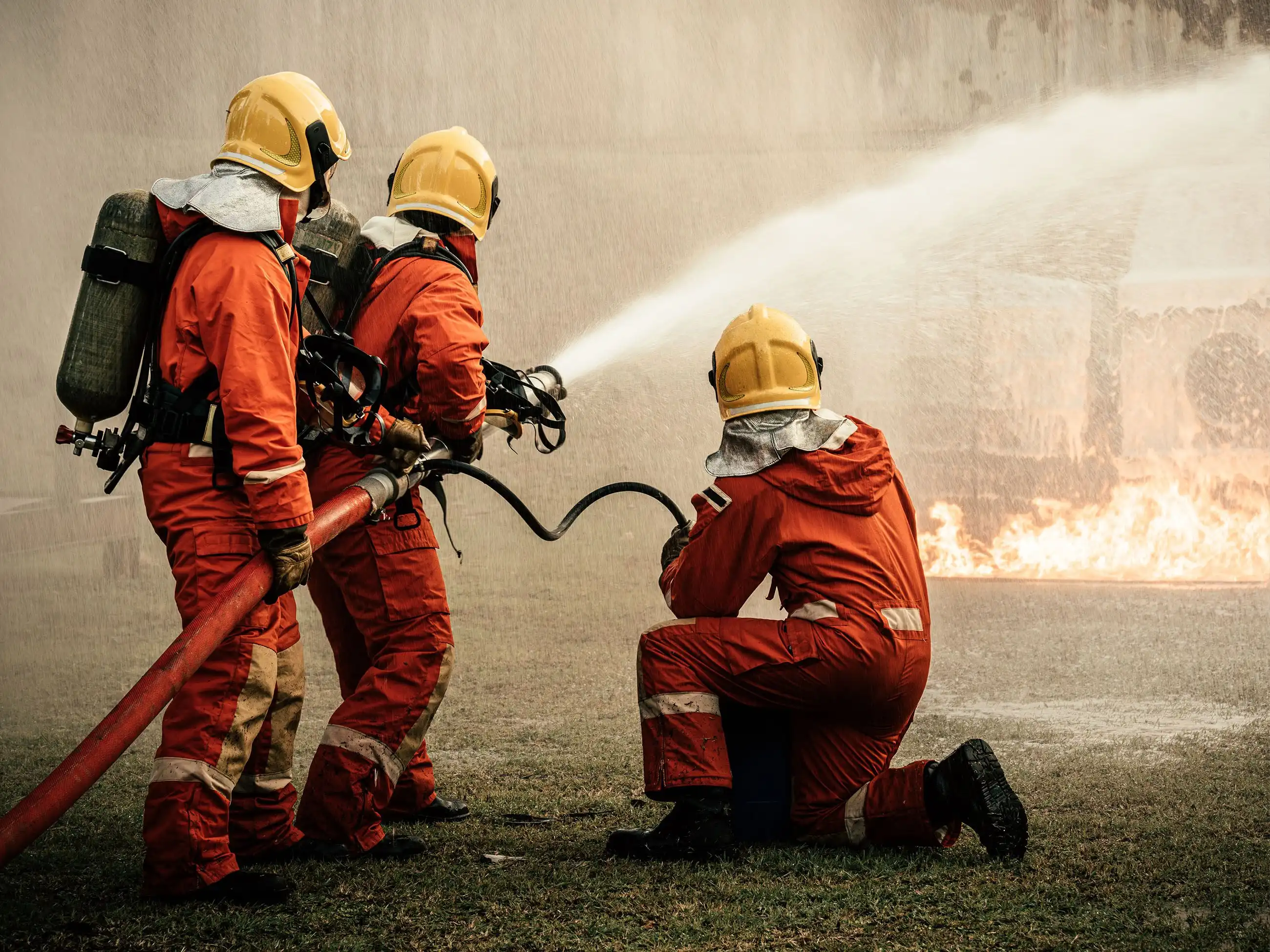 Drei Feuerwehrleute halten einen Wasserschlauch, mit dem sie ein Feuerwehr im Hintergrund des Bildes löschen