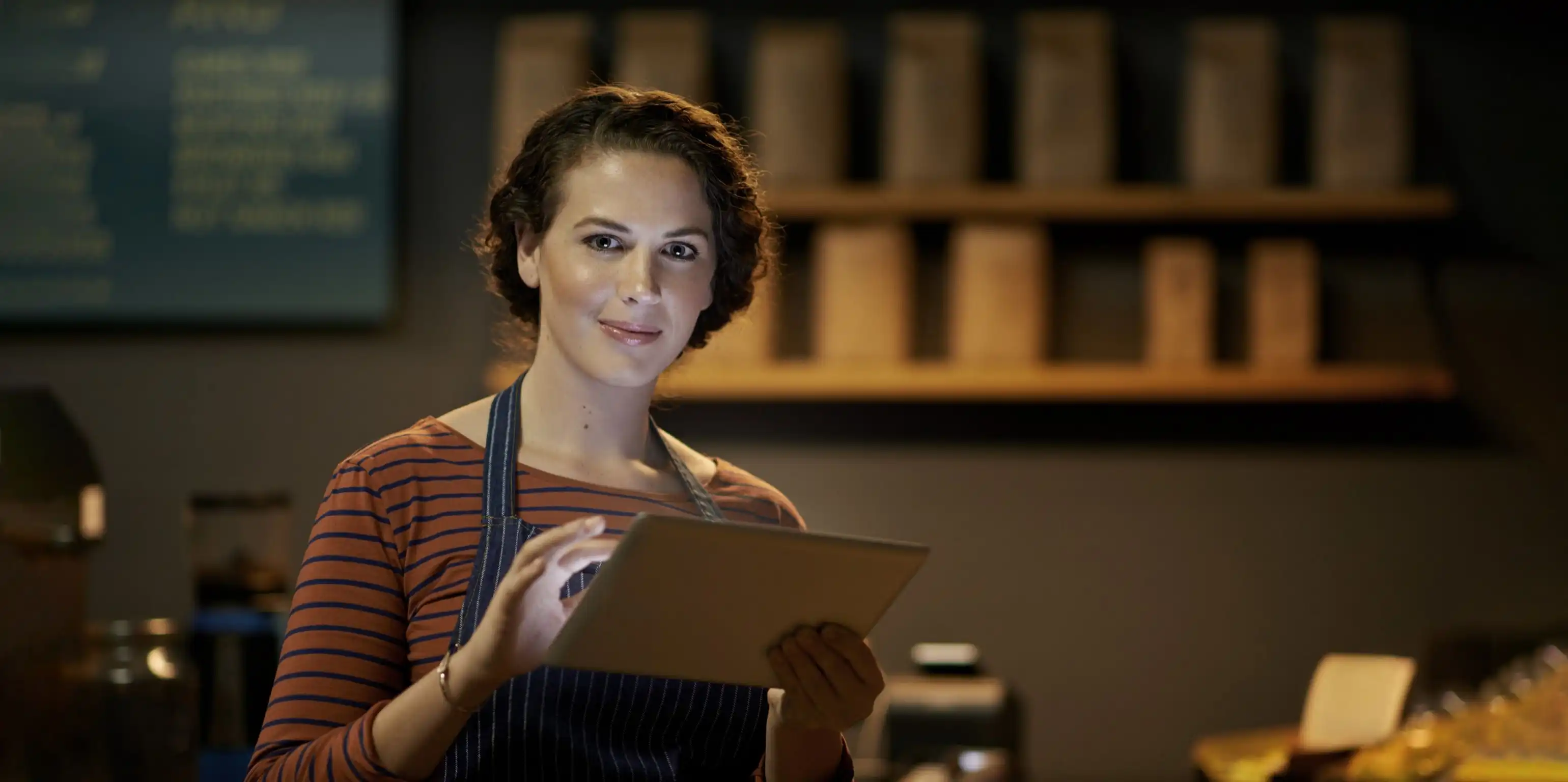 Eine Frau mit Schürze hält ein Tablet in der Hand und steht hinter der Theke eines Cafés