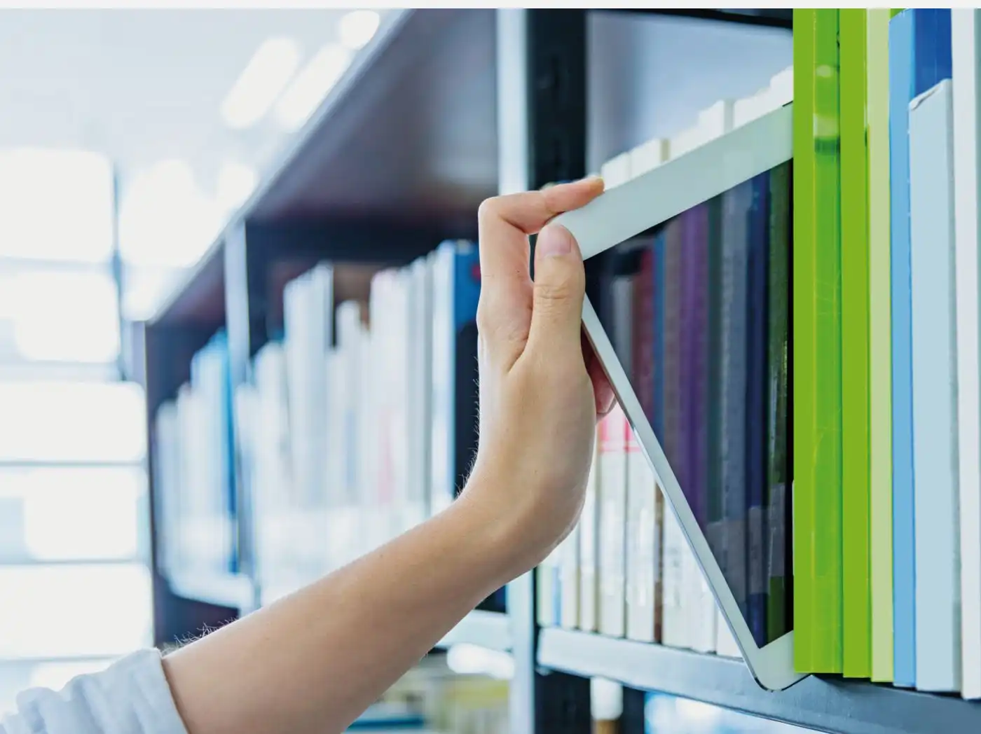 A hand reaches for a tablet placed among numerous books in a bookshelf, in the background many more shelves with books are visible