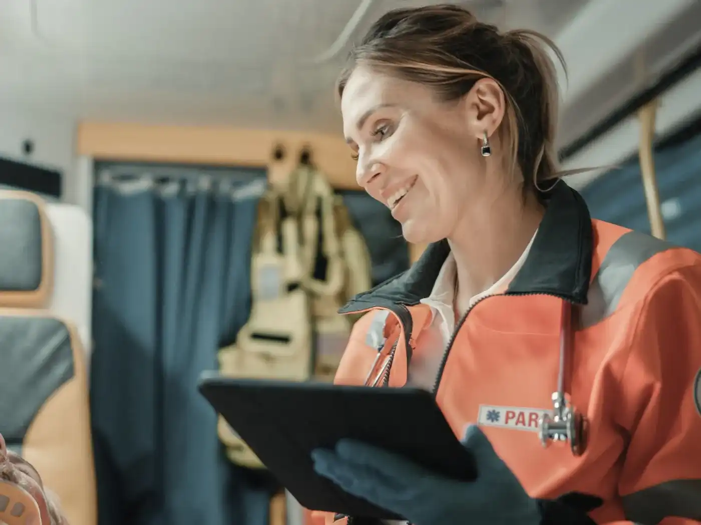 Lächelnde Frau in oranger Rettungsdienst-Jacke mit Stethoskop blickt im Innenraum eines Rettungswagens auf ein Tablet.