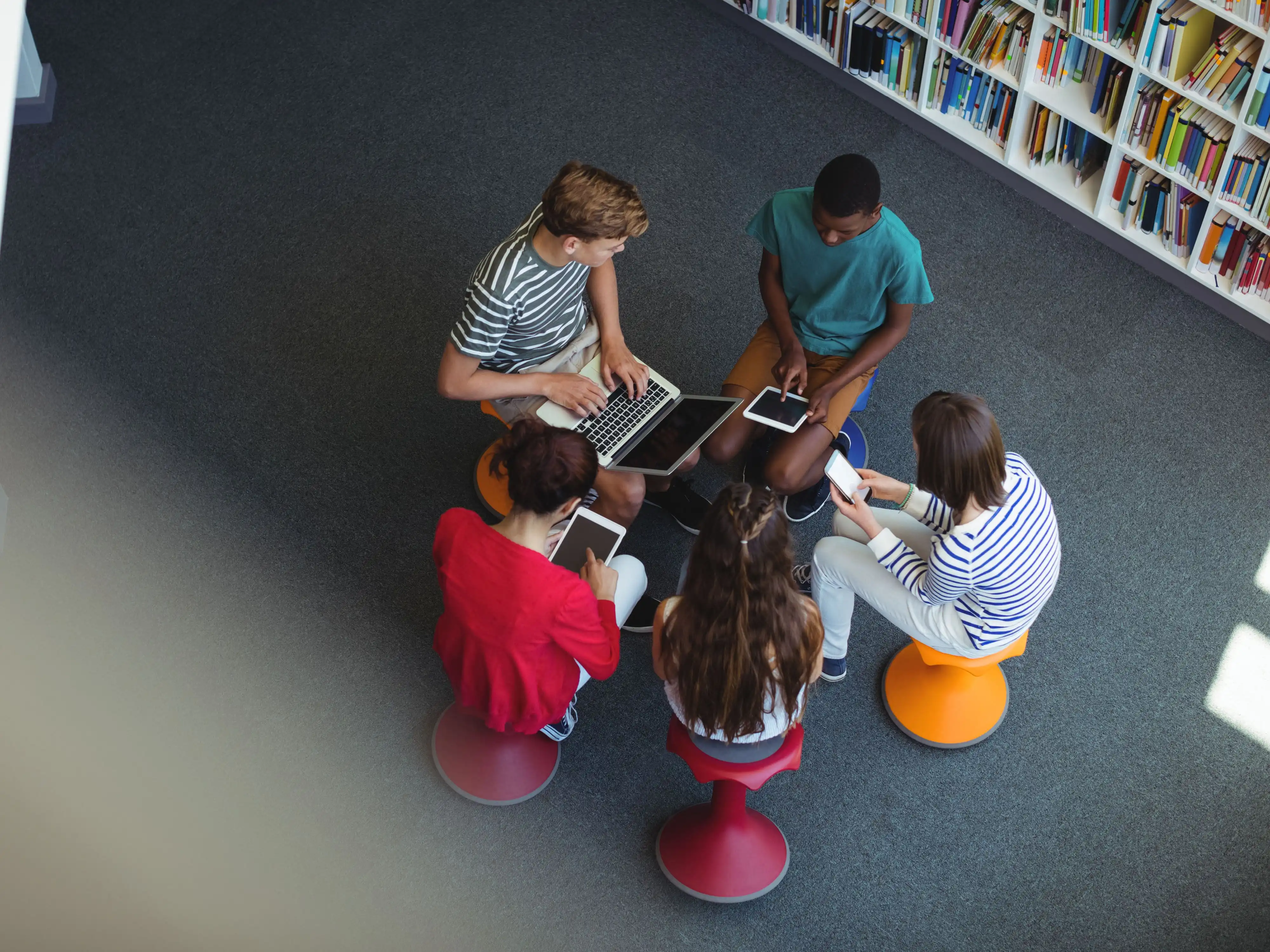 Un groupe de jeunes personnes assises en cercle, tenant chacune un appareil mobile dans la main, avec de nombreuses étagères de livres en arrière-plan