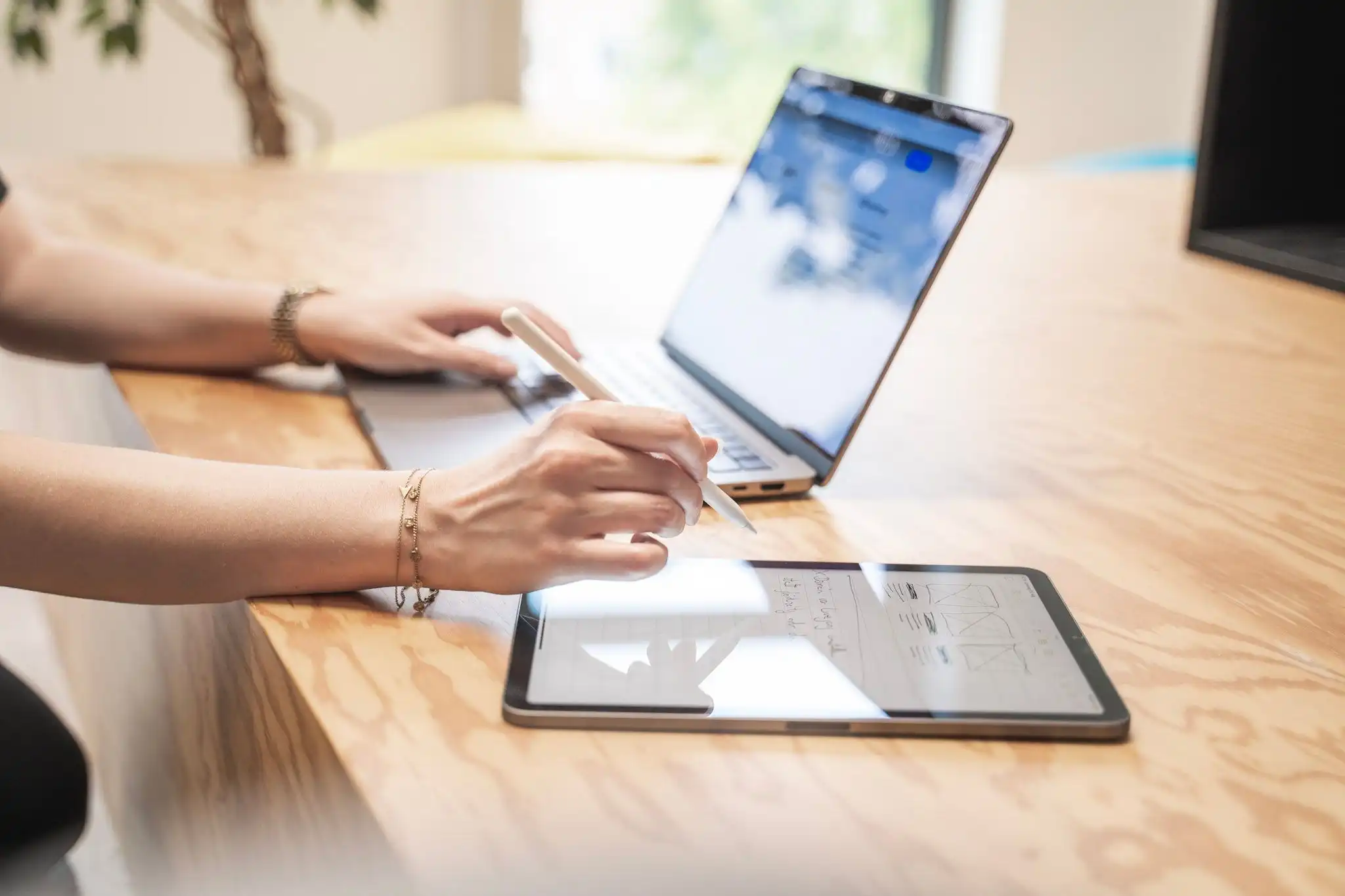 Eine Person arbeitet gleichzeitig an einem Laptop und einem Tablet, beide Geräte stehen auf einem Holztisch, zu sehen sind nur Arme und Hände