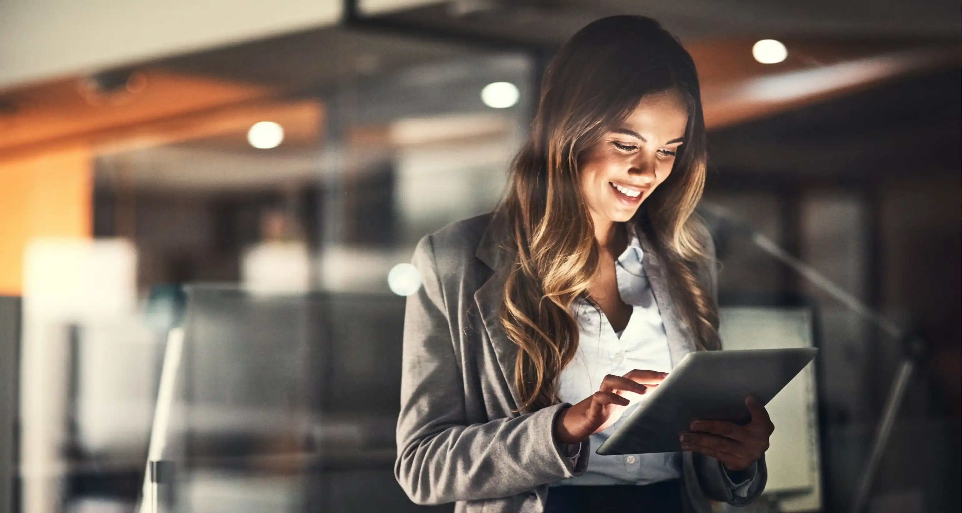 Una donna in abbigliamento professionale tiene un tablet in mano e lo guarda sorridendo, con un ufficio moderno visibile sullo sfondo
