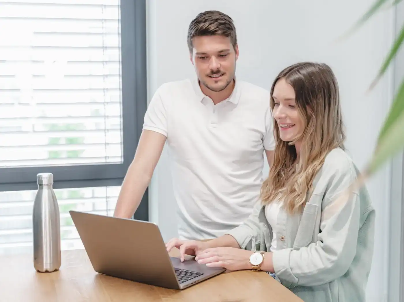 Un hombre y una mujer están de pie frente a un portátil, mirando juntos su pantalla, mientras la mujer tiene su dedo sobre el touchpad