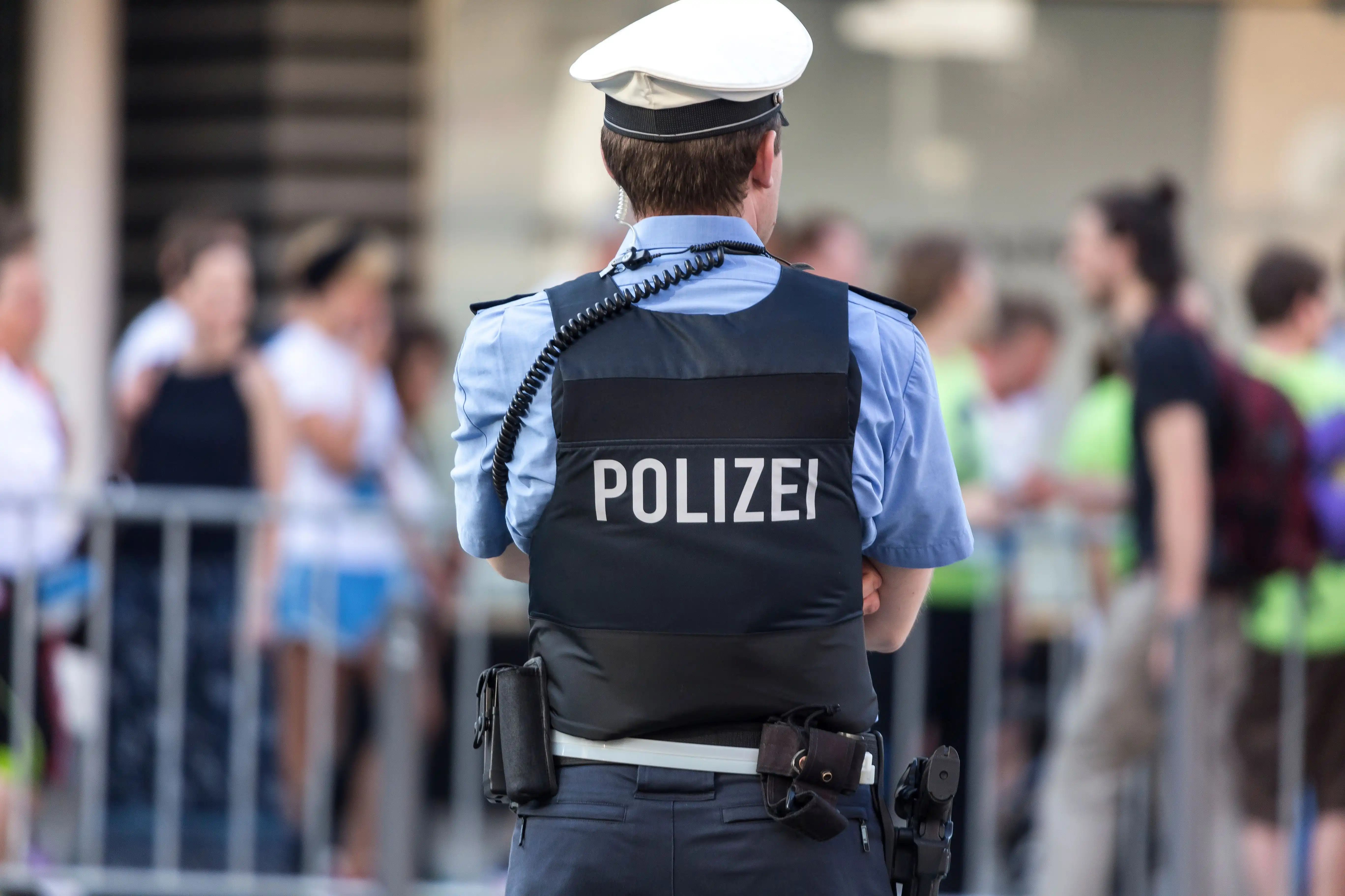 Einzelner Polizeibeamter von hinten mit Schutzweste ‚POLIZEI' und weißer Mütze, im Hintergrund Absperrgitter und eine Menschenmenge.