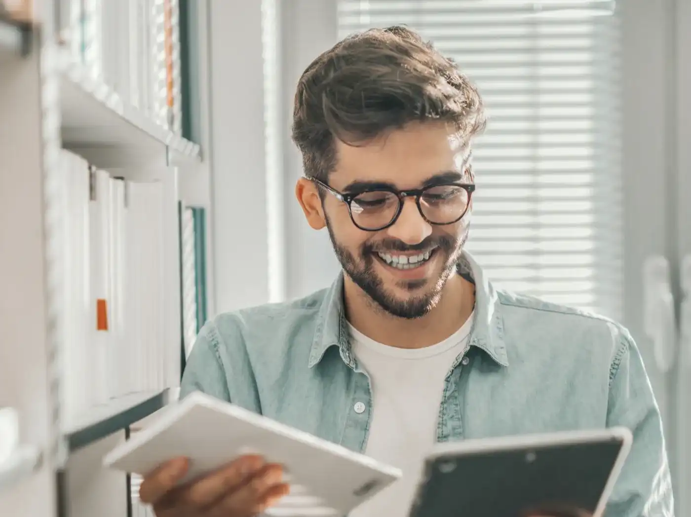 Ein lächelnder Mann mit Brille hält ein Heft und ein Tablet, neben ihm ein Regal mit zahlreichen Büchern