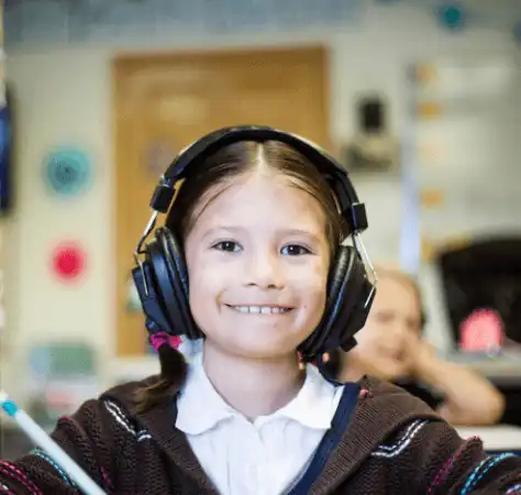 A young girl smiles at the camera while wearing large headphones on her head and holding a pen in her hand, with a classroom-like setting visible in the background