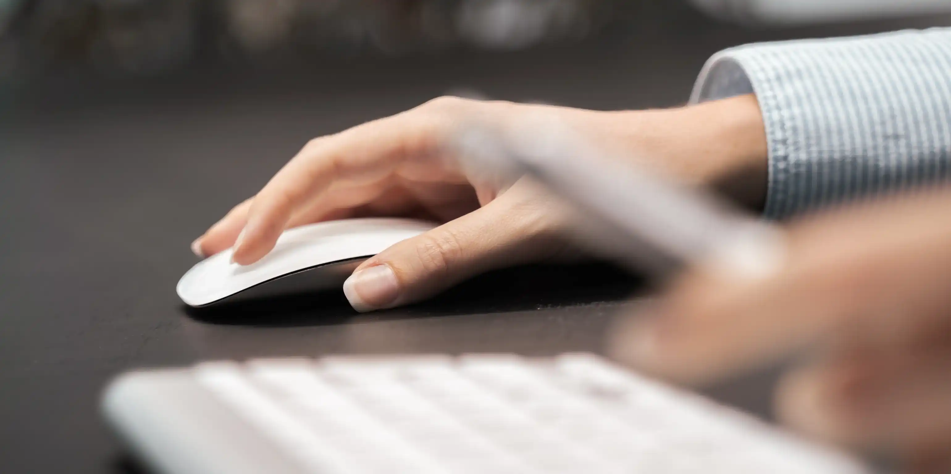 Eine Hand nutzt eine Computermaus, im Vordergrund liegt eine Tastatur, dahinter ist unscharf eine weitere Hand zu sehen