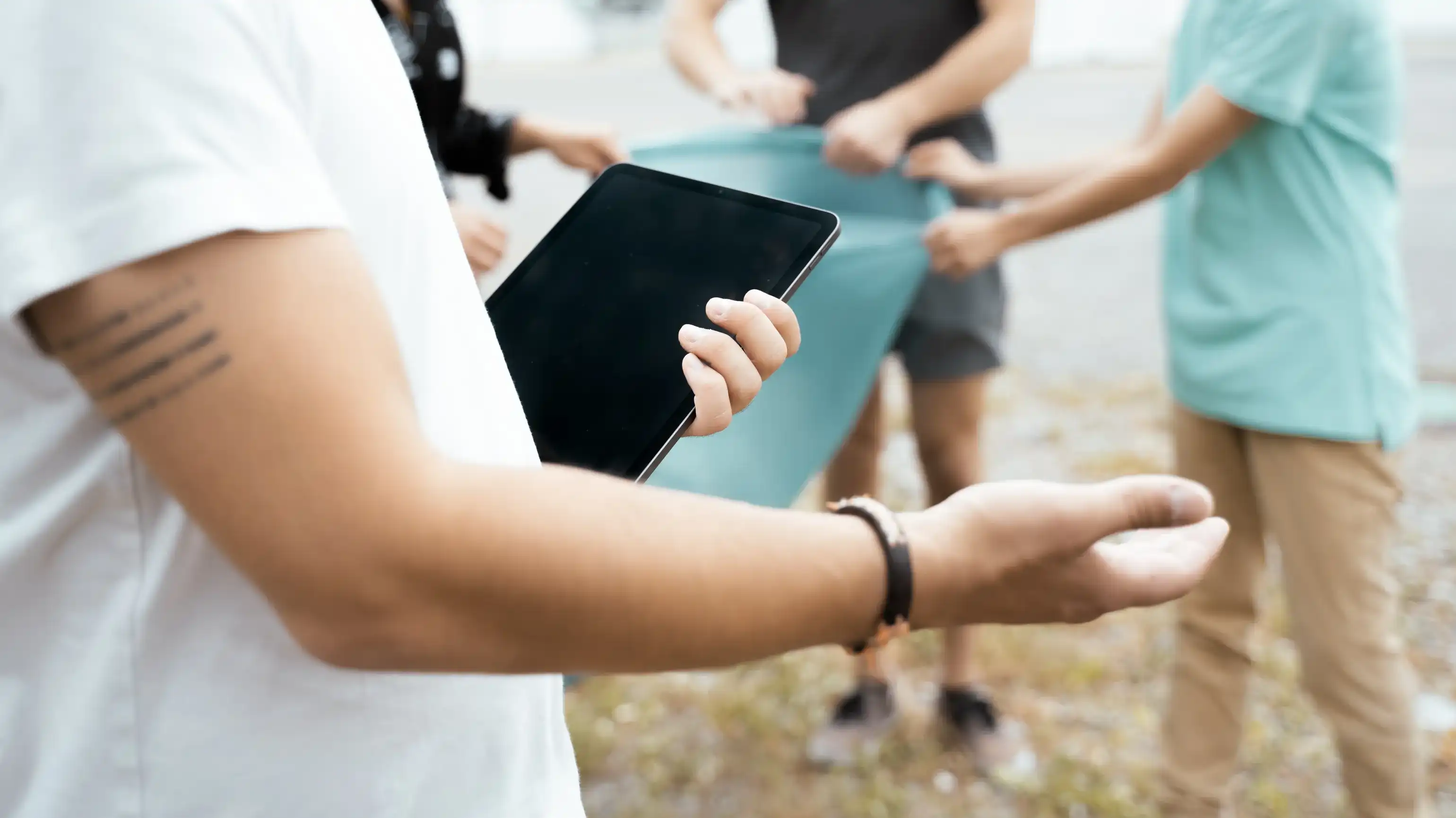 Eine Person hält im Vordergrund ein Tablet, im Hintergrund packen mehrere Menschen im Freien gemeinsam an