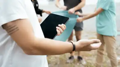 Eine Person hält im Vordergrund ein Tablet, im Hintergrund packen mehrere Menschen im Freien gemeinsam an