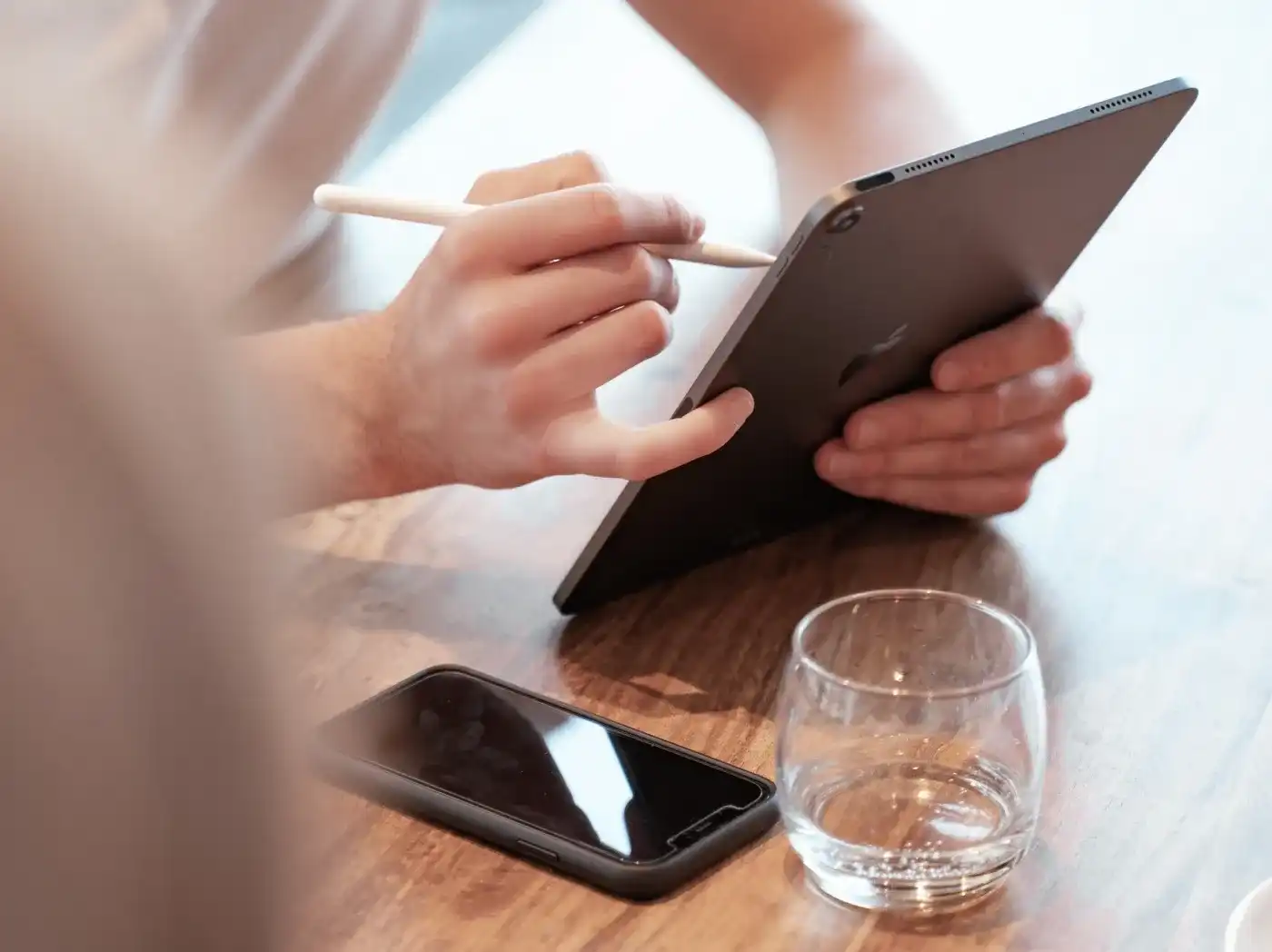 Eine Person arbeitet mit einem Apple Pencil auf einem iPad, neben ihr auf dem Tisch liegt ein iPhone