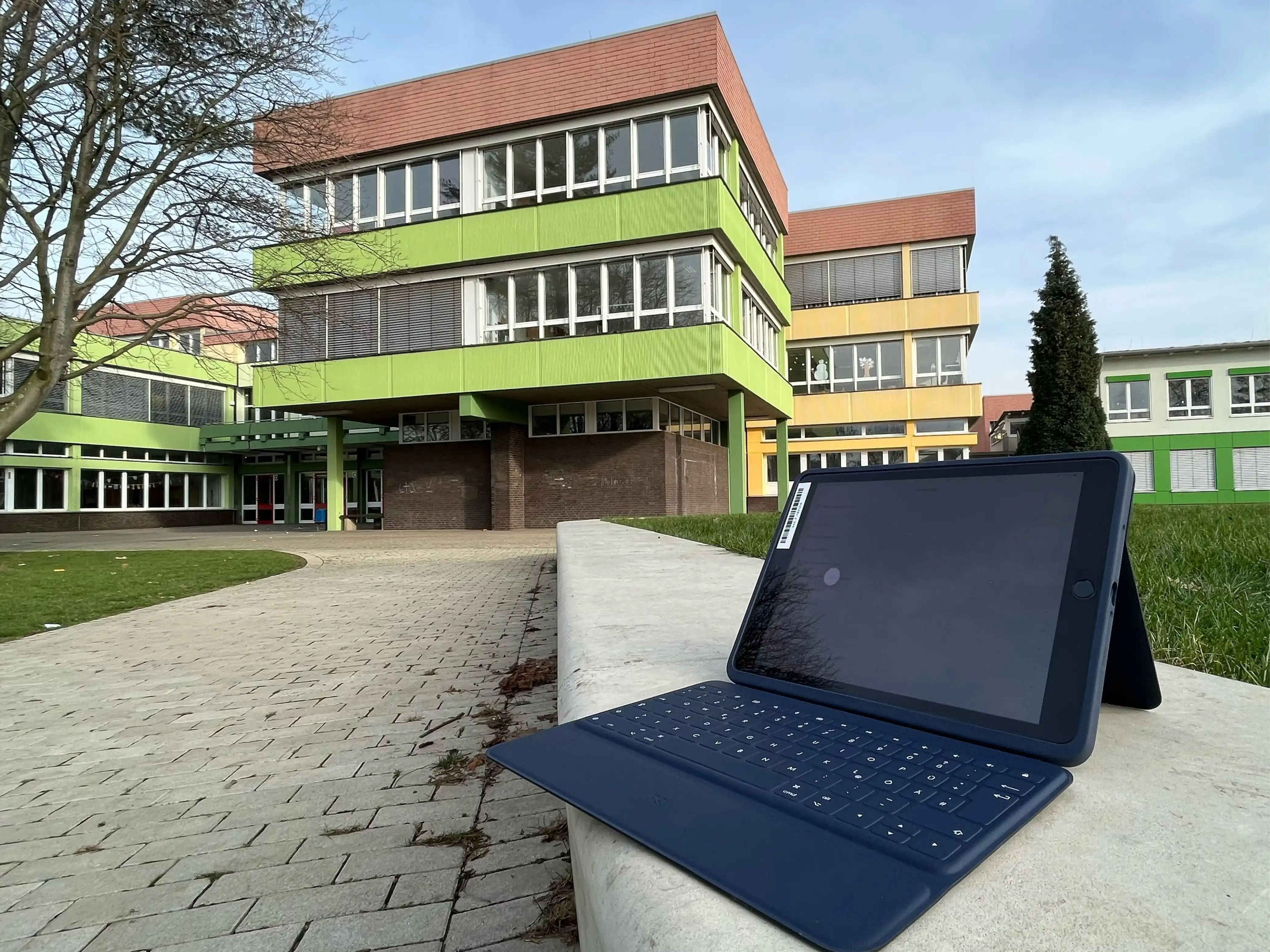 Ein Tablet mit Tastatur steht auf eine Art Steinbank, im Hintergrund ist ein größerer Gebäudekomplex zu sehen, der in bunten Farben gestrichen ist