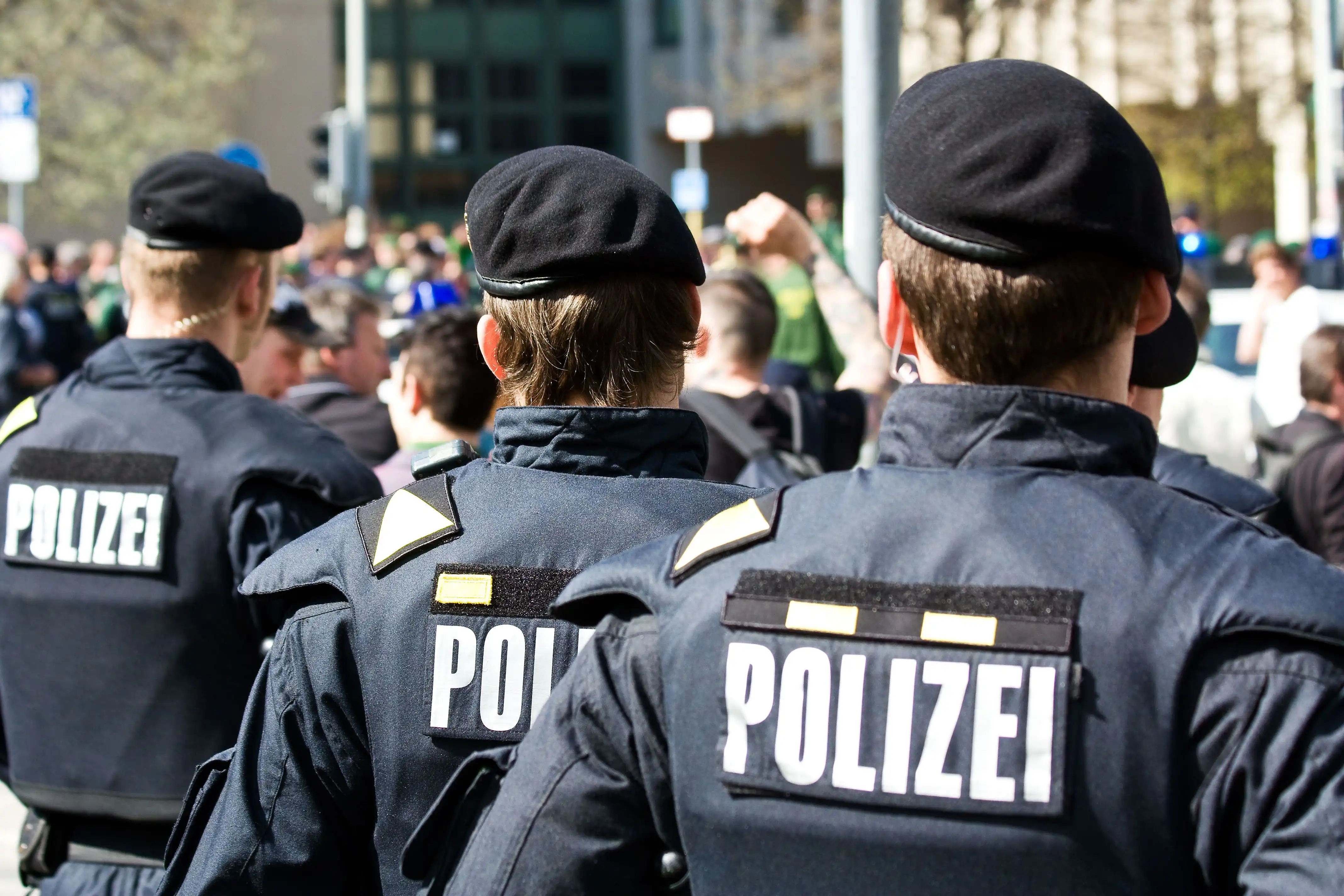 Drei Polizeibeamte von hinten mit Polizei-Westen und Baretten, vor einer Menschenmenge im Freien.