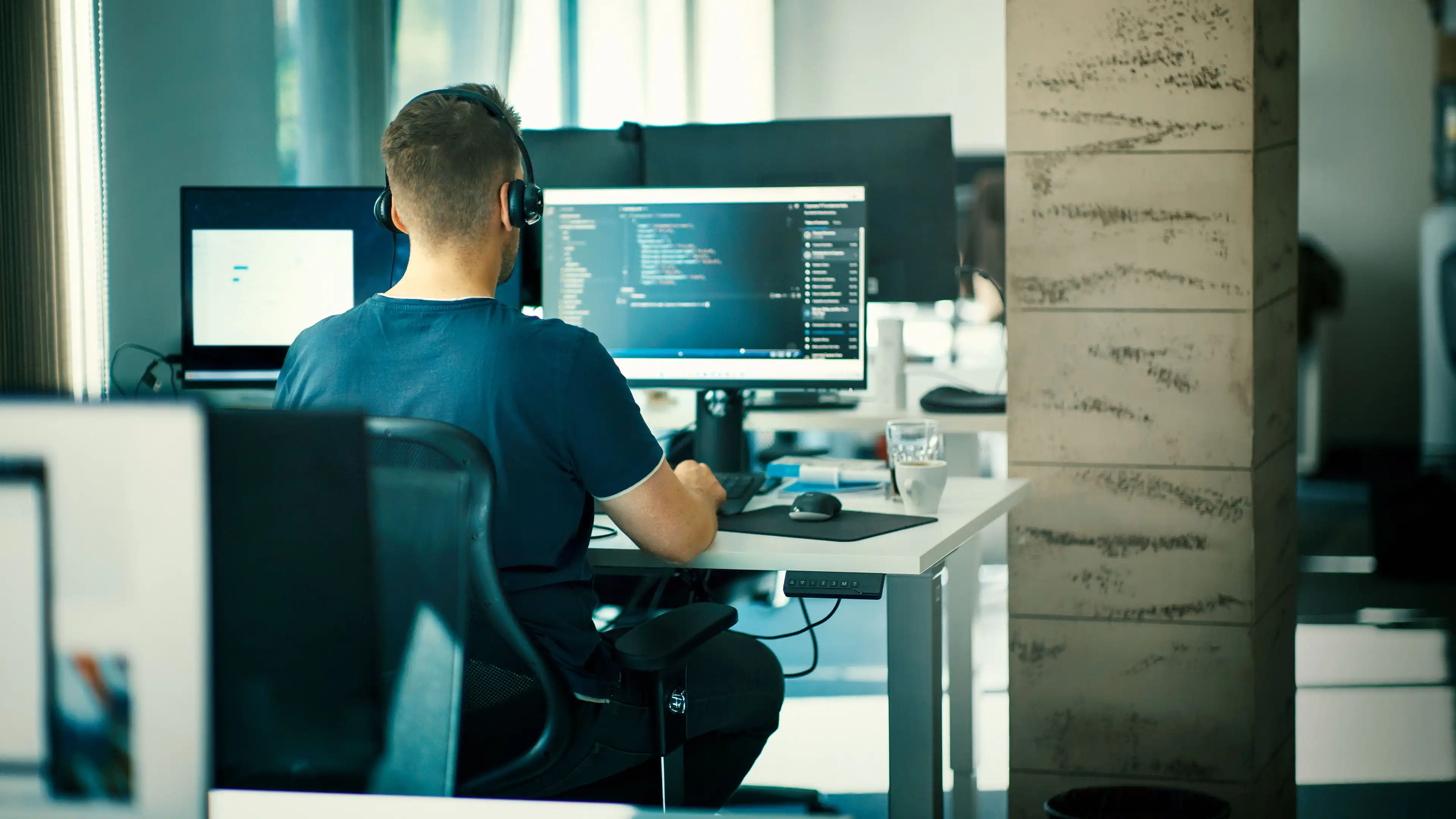 A person wearing headphones, seen from behind at a desk in an open-plan office, in front of a monitor displaying program code and a laptop