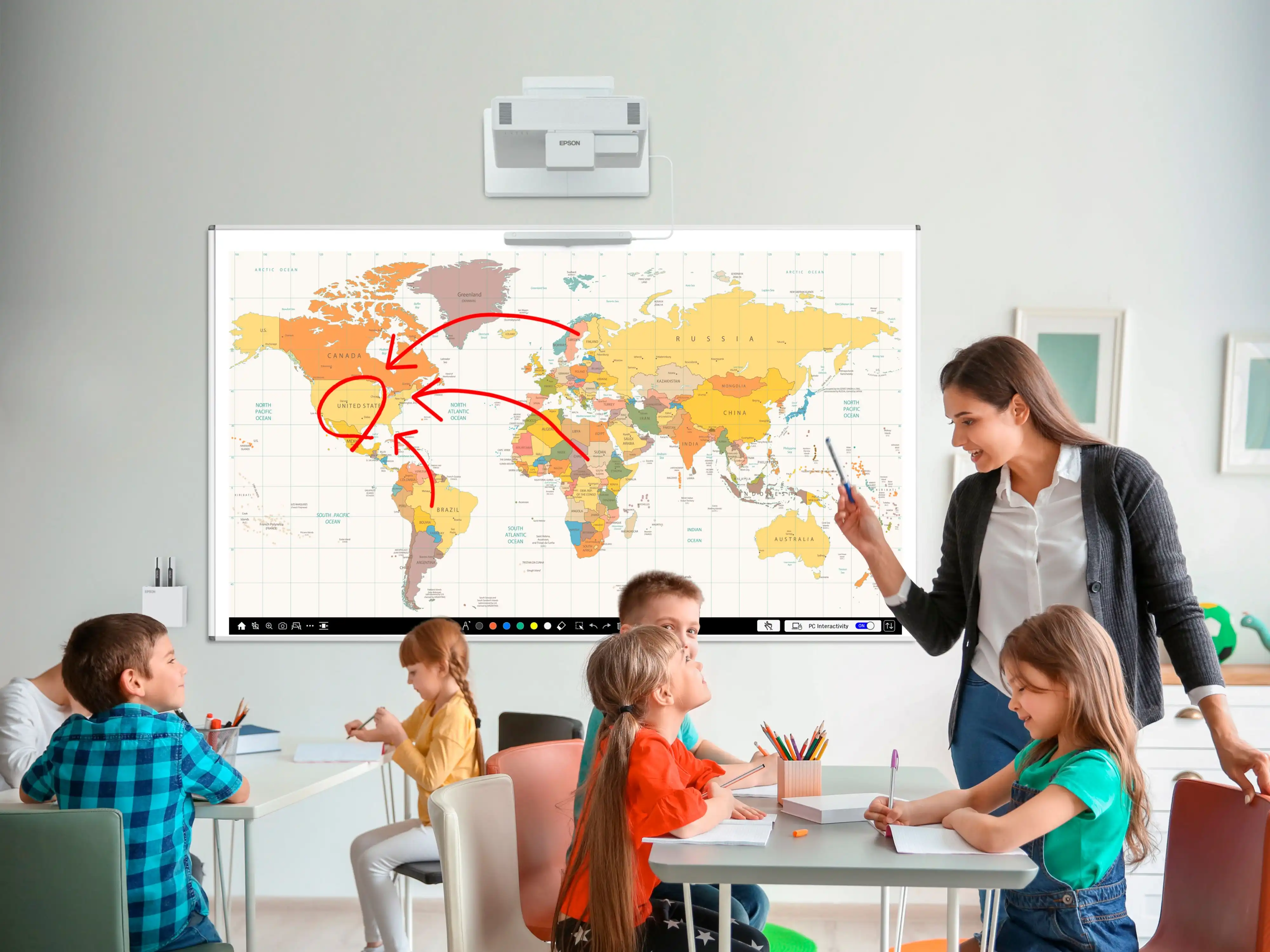 Eine Frau zeigt mit einem Stift auf ein interaktives Whiteboard mit Weltkarte, über dem ein Epson-Projektor hängt, während Kinder an Tischen davor sitzen