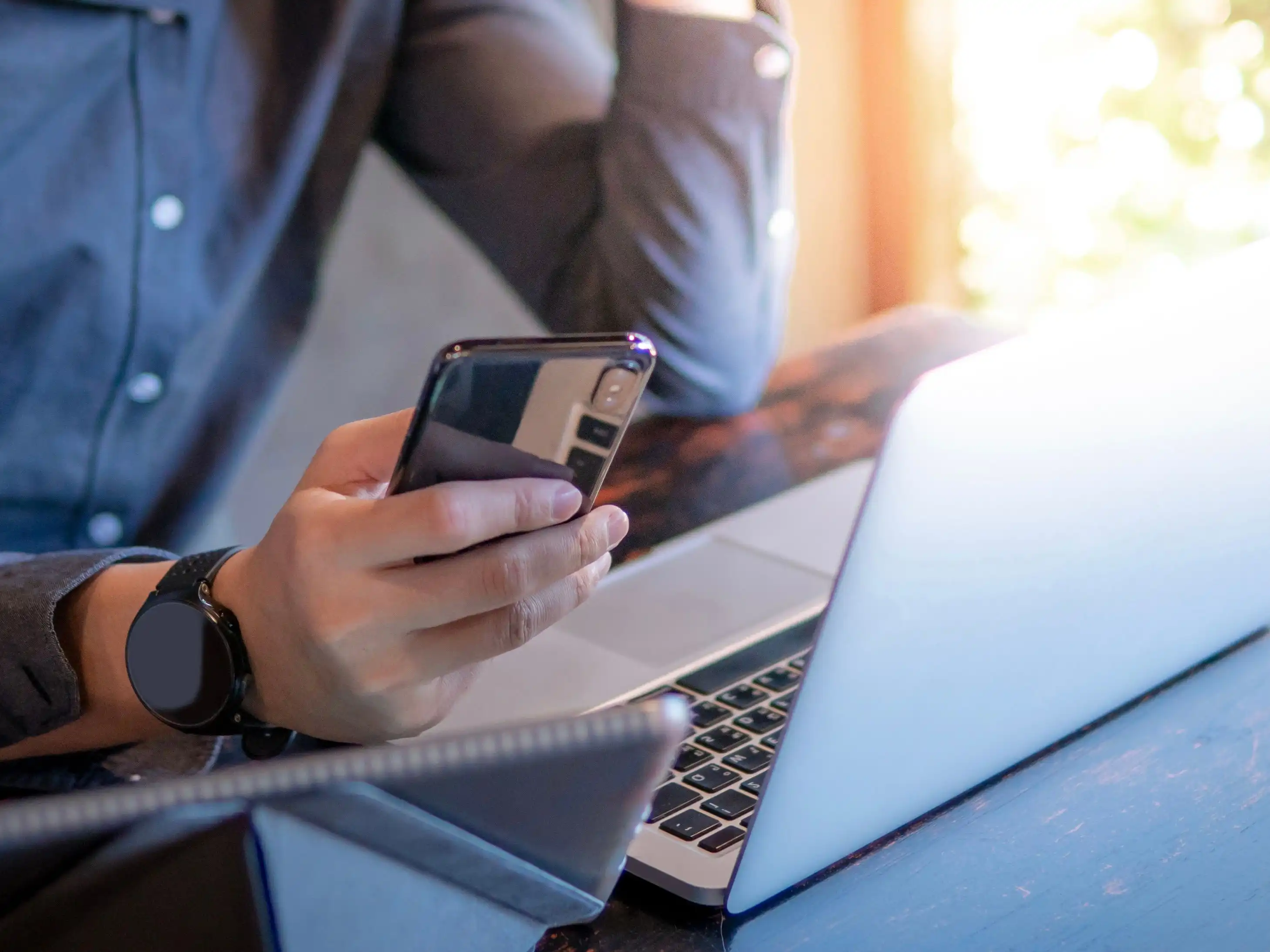Ein Mann in einem Hemd hält ein Handy in der Hand und sitzt vor einem Laptop.