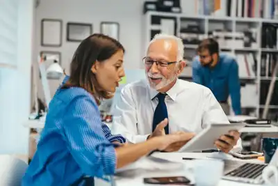 Eine jüngere Frau und ein älterer Mann mit Brille sitzen in einem Büro und schauen gemeinsam auf ein Tablet, im Hintergrund arbeitet eine weitere Person