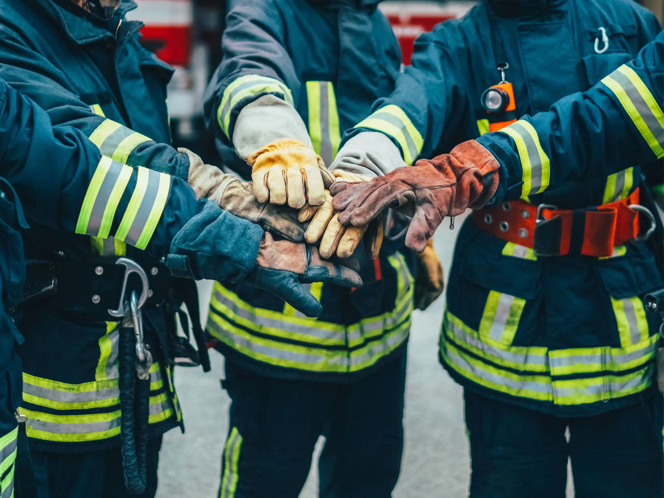 Mehrere Feuerwehrleute in voller Ausstattung legen ihre Hände, die entsprechende Handschuhe tragen, aufeinander, als Symbolbild für Zusammenarbeit